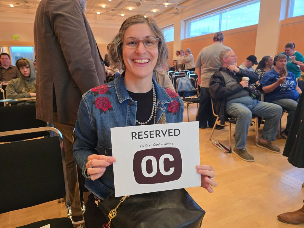A smiling woman holding a sign that reads 'RESERVED For Open Caption Viewing OC' while seated in a crowded room.