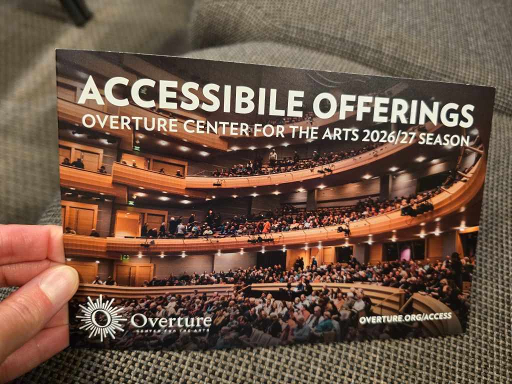 A hand holding a promotional card for accessible offerings at the Overture Center for the Arts, featuring a packed theater with multiple levels and audience members.